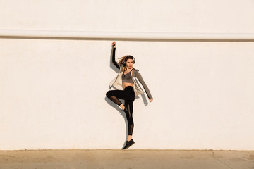 Full length shot of young pretty healthy woman in sport wear jumping near white wall