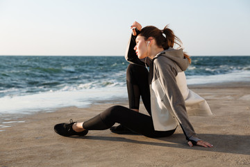 Photo of charming sport woman sitting on the beach wipes the sweat from head while resting after run