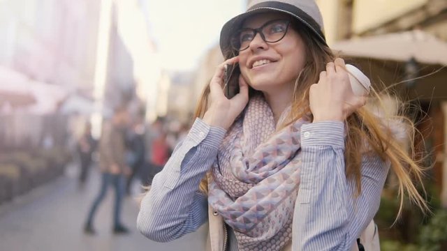 Joyful caucasian girl, in striped hat and coloured scarf, chatting on phone while walking in the street, sunny day, slowmotion