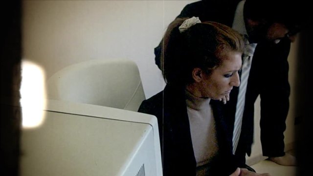 Fake 8mm amateur film: a male employee, standing still, annoys his female coworker, asks stupid questions, takes her keyboard, makes a kind of mobbing.

