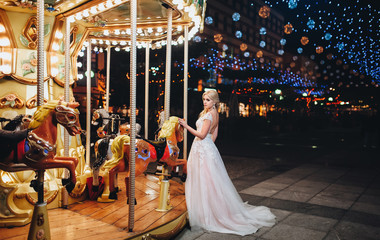 The bride blond in a light pink wedding dress is standing near the carousel in the background of a night cityscape . 