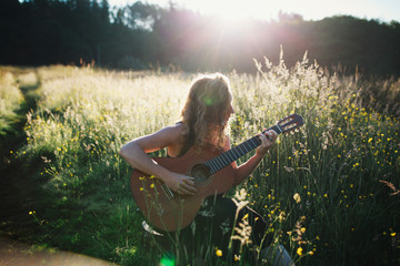Young woman sitting in field at sunrise playing classical guitar and singing