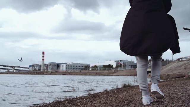Unrecognisable person in winter streetwear parket walking by water shore covered in rocks at urban city area on cold cloudy day