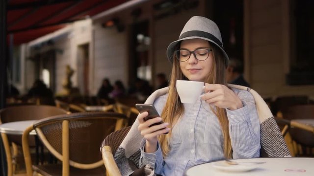 Smiling girl, in a grey striped hat and glasses, using mobile phone during coffee time in the outside cafe, slowmotion