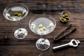 Make martini cocktails. Glass with beverage, olives and utensils on dark wooden background top view