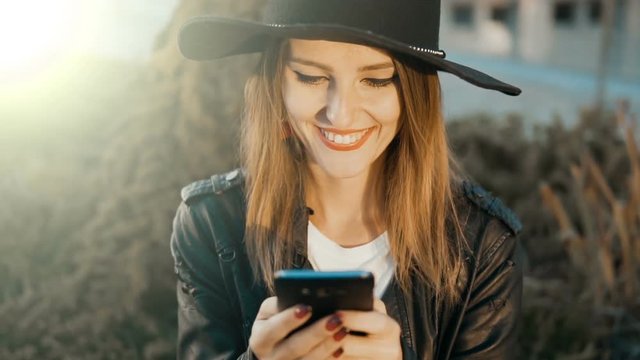 Joyful caucasian girl, in black hat and leather jacket, texting on the phone, outdoor slowmotion