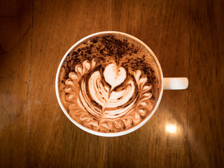 Cappuccino with fresh milk foam and latte art in a white ceramic glass on a wooden table at a coffee shop.