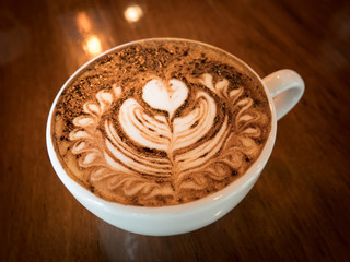 Cappuccino with fresh milk foam and latte art in a white ceramic glass on a wooden table at a coffee shop.