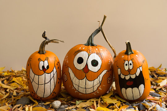 Three smiling and laughing pumpkins with painted faces that have oval eyes and big teeth. The pumpkins are photographed against a beige background..