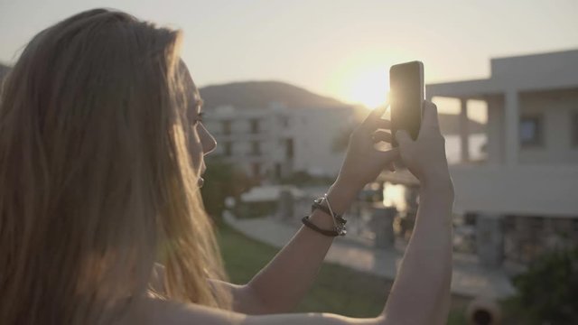 Young blonde woman is taking photo with smartphone and looking at panorama view of resort at sunset. Attractive girl takes pictures on cell phone. Vacation and destination concept.