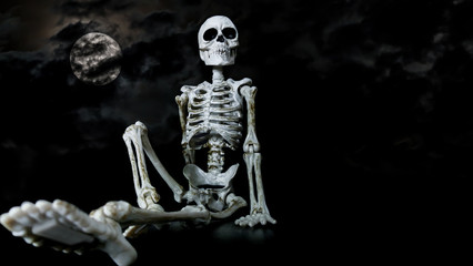Halloween toy skeleton sitting down in a relaxed manner with the moon and dark clouds behind.
