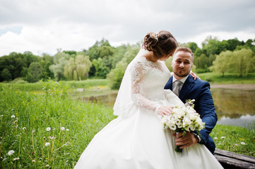 Fantastic bride sitting on a groom's lap in the meadow next to the lake on a wedding day.