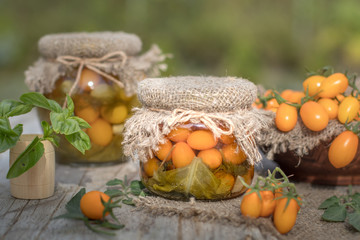Preserved autumn vegetables for winter. Delicious homemade canned marinated orange yellow cherry tomatoes with herbs glass jar burlap decoration Rustic style wooden table background