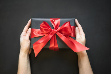 Gift box wrapped in black paper with red ribbon in female hand on black surface.