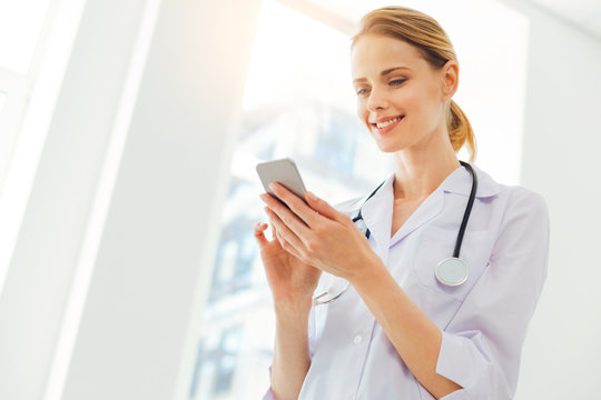 Joyful young woman in labcoat using smartphone at clinic