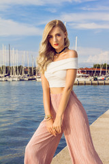 Beautiful young woman with long curly blonde hair with a beautiful makeup posing on the harbor. In the background are beautiful clouds. Around her neck is a stylish black choker.
