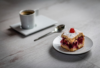Rectangular slice of fruit cake with a strawberry on top on a round plate and a round cup of espresso coffee on a rectangular plate with a tea spoon in the middle