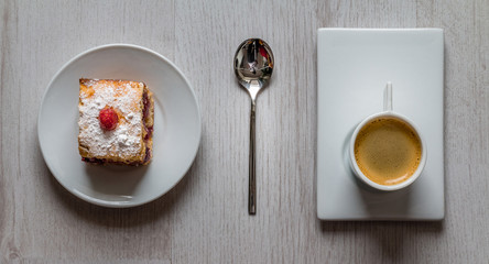 Rectangular slice of fruit cake with a strawberry on top on a round plate and a round cup of espresso coffee on a rectangular plate with a tea spoon in the middle