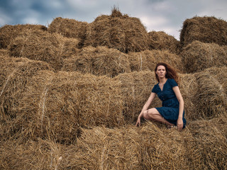 Naklejka na ściany i meble Red-haired girl in a light summer dress of blue color sits on a stack of straw.