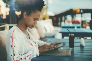 Serious black lady in traditional African chemise is sitting at the teal wooden table of street cafeteria and working on digital tablet while waiting for her friend, with glass of beverage near