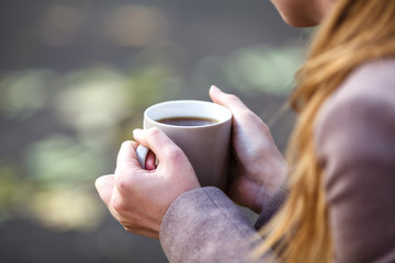 Closeup view of disposable cup of coffee or tea in womans hand. Autumn park with trees and yellow leafes