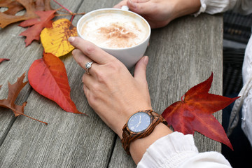 Woman with elegant hands wearing a wooden watch holding a cup of coffee against a rustic table with colorful autumn leaves
