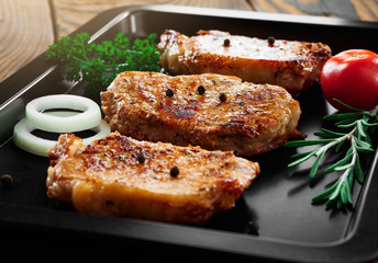 Baked pork steaks on a black baking sheet on wooden background