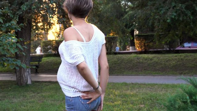 Lovely pregnant girl in a city park at sunset. She puts on sunglasses and strokes the belly