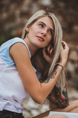Naklejka premium Beautiful blonde woman sitting on a stone embankment and reads book. Hands tattooed. modern, trendy, student. Closeup portrait