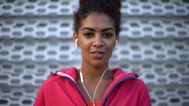 picture of happy woman with tied dark hair wearing sport clothes enjoying music with closed eyes listening via headphones looking at camera with grey wall background slow motion