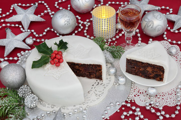 Iced christmas cake and slice with glass of sherry, holly, fir, bauble decorations and silver foil wrapped chocolates on red background.