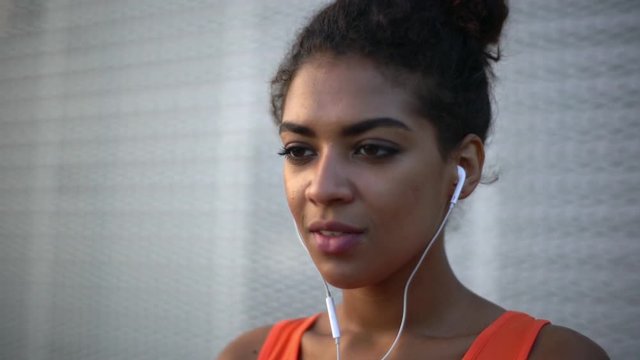 closeup picture of sporty young afro-american woman in bright shirt putting on headphones listening favourite music looking ahead with grey wall background slow motion