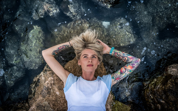 Portrait of a beautiful blonde which lies on the stone near the sea. Women's hands with a tattoo. Modern fashion girl