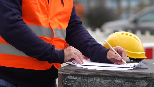 Cropped image of a left-handed construction worker wearing protective clothing taking notes on construction site