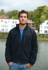 junger Mann mit Jacke am Fluss vor Neubauten,Blick in die Weite oder ins Leere