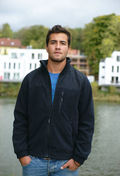 junger Mann mit Jacke am Fluss vor Neubauten,Blick in die Weite oder ins Leere