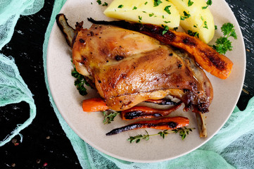 Fried rabbit leg, garnish of boiled potatoes, grilled carrots - on a plate on a dark background. Top view.