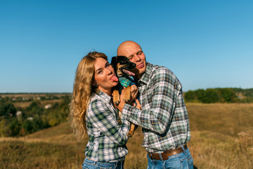 male guy in checkered shirt and girl in green dressed in a black shirt and jeans kiss standing in grass sitting in grass half on top of sunset with dog running around fooling around