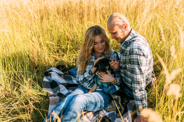 Naklejka na ściany i meble male guy in checkered shirt and girl in green dressed in a black shirt and jeans kiss standing in grass sitting in grass half on top of sunset with dog running around fooling around