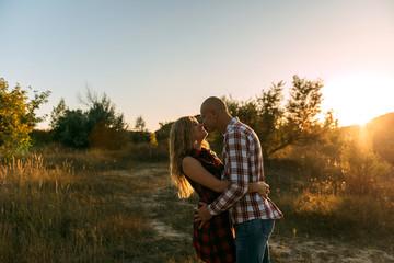 man guy in plaid shirt and woman girl in red checkered dress kiss standing in grass sitting in grass half on top on sunset background