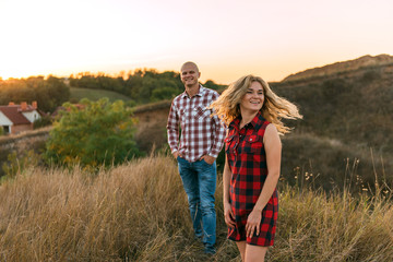guy man in plaid shirt and girl in red checkered dress kiss standing in grass half top on sunset background