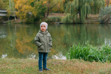 Fototapeta na wymiar boy child in a green jacket and a gray hat smiles against the background of the autumn forest of the park of the lake