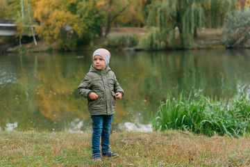 Fototapeta na wymiar boy child in a green jacket and a gray hat smiles against the background of the autumn forest of the park of the lake