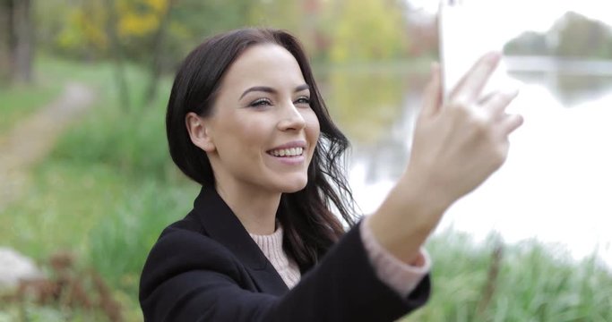 Beautiful woman in black jacket and polo-neck shirt taking selfie near pond during her walk in autumn park.