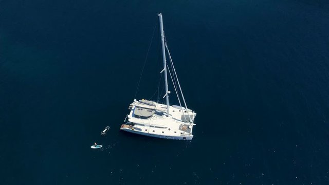 Aerial Top Down Shot of a Big White  Sailing Yacht Anchored in a Bay with Man Standup Paddleboarding Near it. Ocean is Dark Blue and Beautiful. Shot on Phantom 4K UHD Camera.