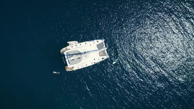 Aerial Top Down Shot of a Big White  Sailing Yacht Anchored in a Bay with Man Standup Paddleboarding Near it. Ocean is Dark Blue and Beautiful. Shot on Phantom 4K UHD Camera.