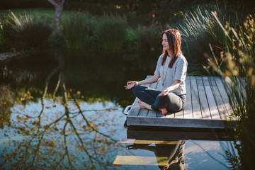 Beautiful young woman meditating on wooden deck at waters edge