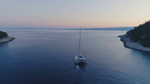 Tranquil Aerial Shot of a Catamaran Yacht Sailing out of the Beautiful Bay. It's Evening Sky is with a Tint of Pink and Tropical Coasts Visible. Shot on Phantom 4K UHD Camera.