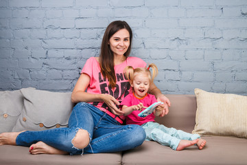 beautiful young mother uses a smartphone to play with a two-year-old daughter sitting on the couch in the room Dressed in bright colors, colored clothes
