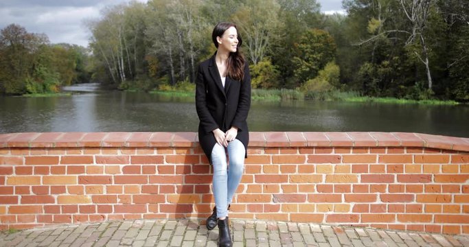 Beautiful woman in black jacket sitting on bridge and enjoying weather during walk in autumn park on sunny day.